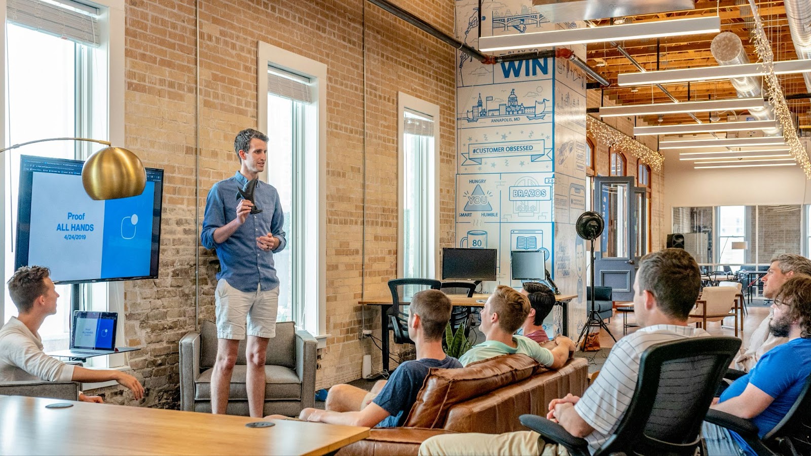 Image shows a sales training session in progress. A man stands at the front to present the training, while trainees sit on sofas and comfortable chairs to listen.